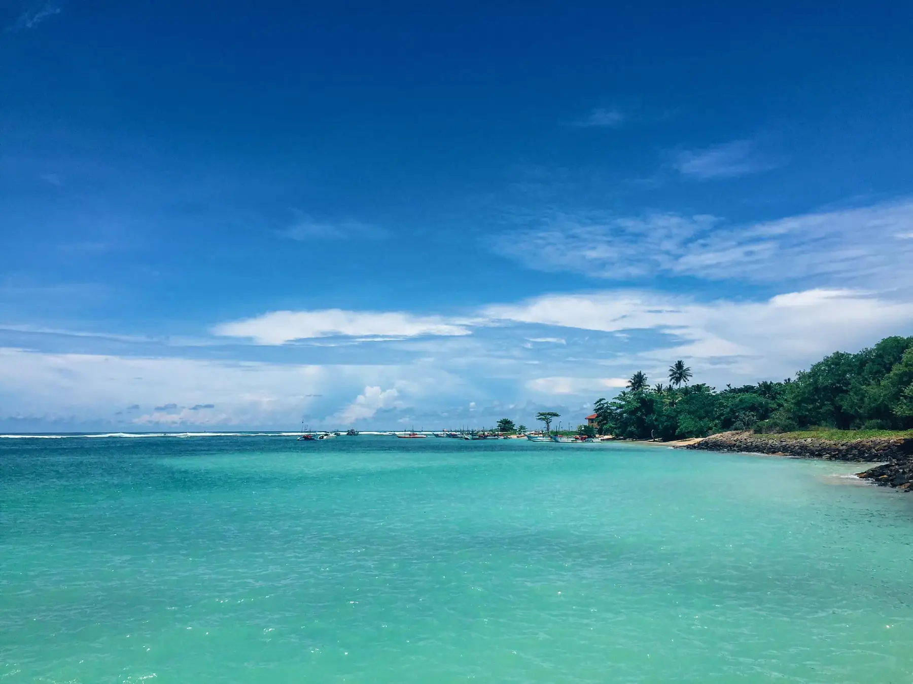 Turquoise water and a palm-lined rocky shore at Ahangama on Sri Lanka's south coast, with fishing boats in the distance.