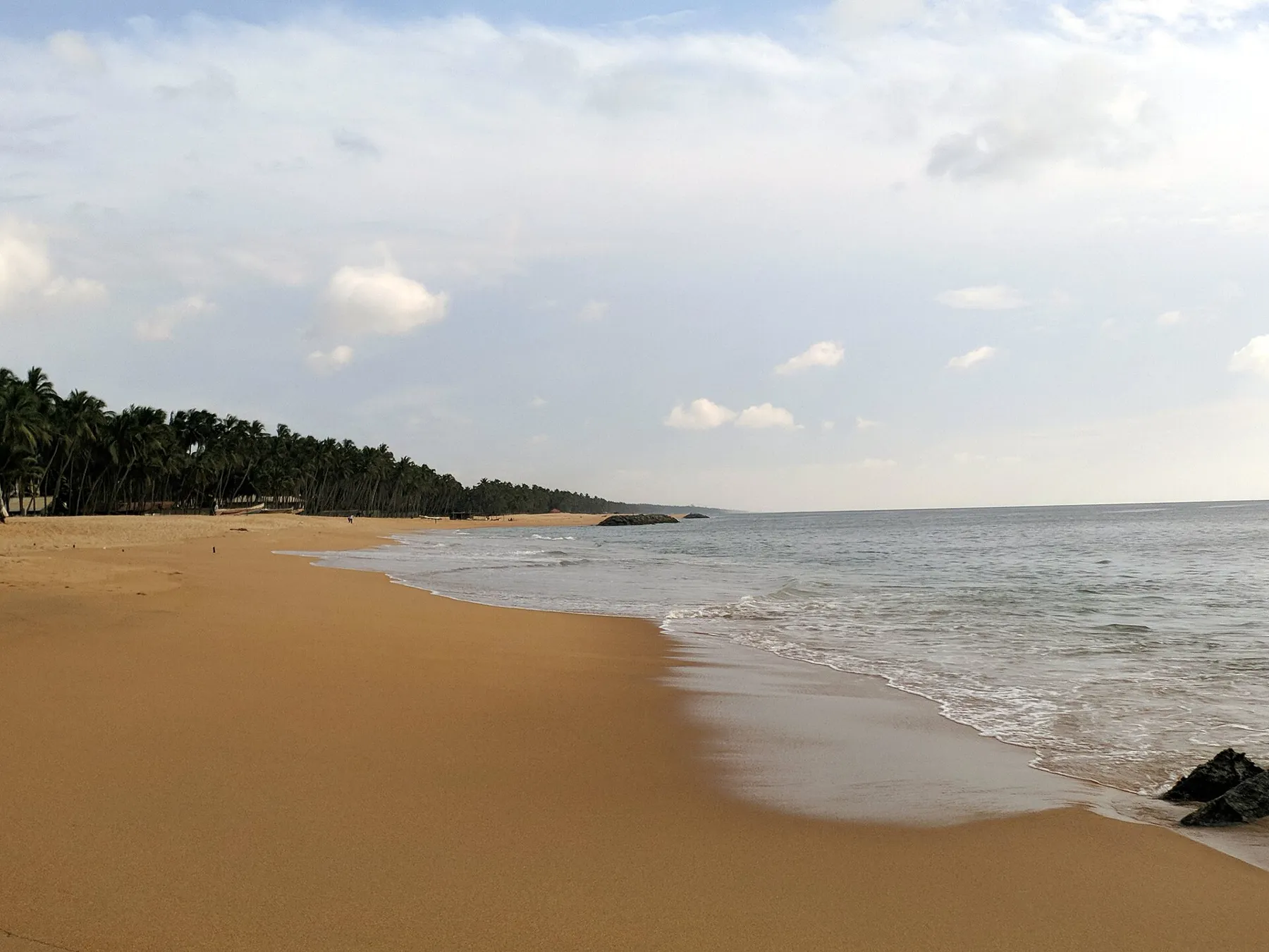 A wide golden beach at Marawila curving toward a line of palm trees, with gentle waves under a partly cloudy sky.