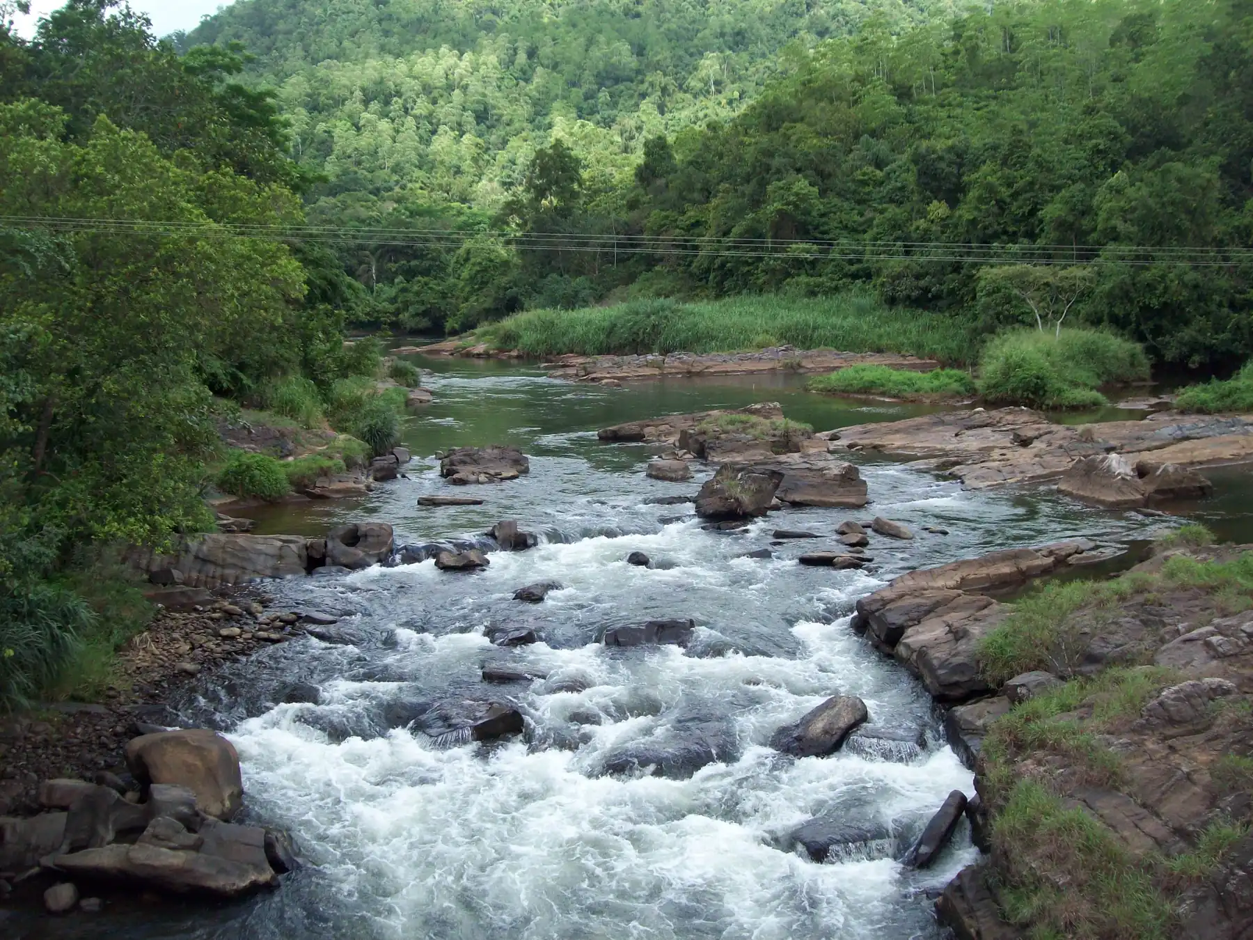 White-water rapids running between dark boulders on the Kelani River at Kitulgala, with forested hills rising behind.